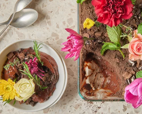 Chocolate dirt pudding with cookie crumble topping garnished with edible flowers in a glass baking dish shown with a serving of pudding in a bowl with spoons