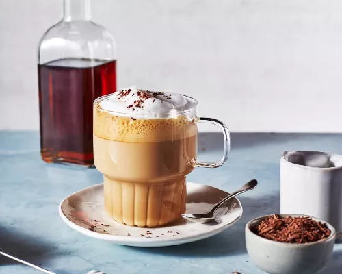 A glass mug full of a frothed latte with whipped topping and shaved chocolate shown with a bottle of brown sugar simple syrup.