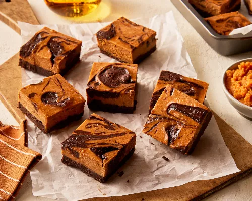 Seven pumpkin cheesecake brownies on a wooden cutting board lined with parchment, shown with a baking pan with brownies and a bowl of pumpkin puree.