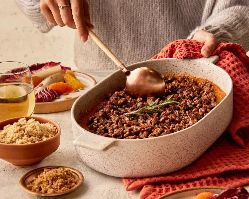 A woman wearing a sweater serving sweet potato casserole garnished with a rosemary sprig from a baking dish, shown with a radicchio and citrus salad, glasses of wine, and bowls of golden yellow and demerara sugar.