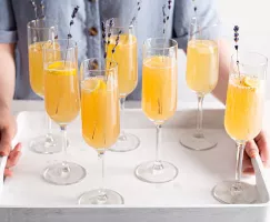 Seven champagne flutes of mimosa on a serving tray held by two hands