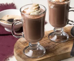 Two mugs of hot chocolate with whipped cream on top, sitting on a wooden board